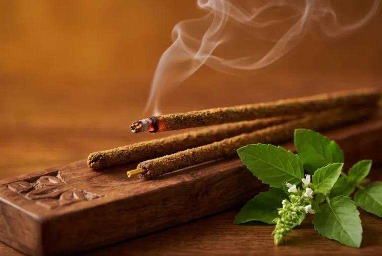 Hand-rolled tulsi incense sticks burning on a wooden incense holder with fresh holy basil leaves and soft smoke